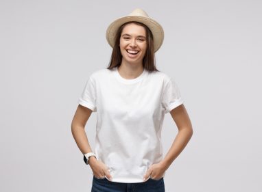 Young smiling european woman dressed in white t-shirt and summer hat, feeling positive and laughing, isolated on gray background